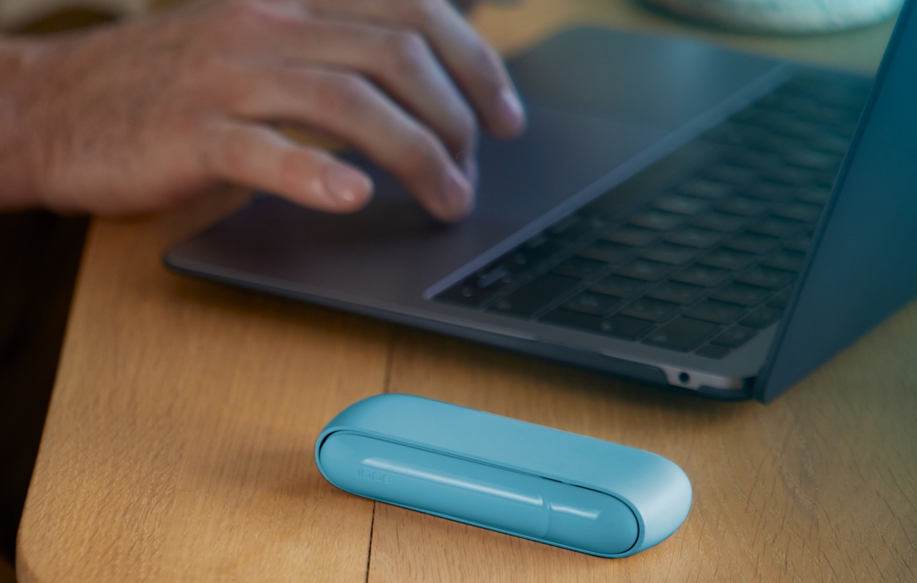 Manos escribiendo en un laptop con un cargador portátil de IQOS Originals DUO turquesa al lado