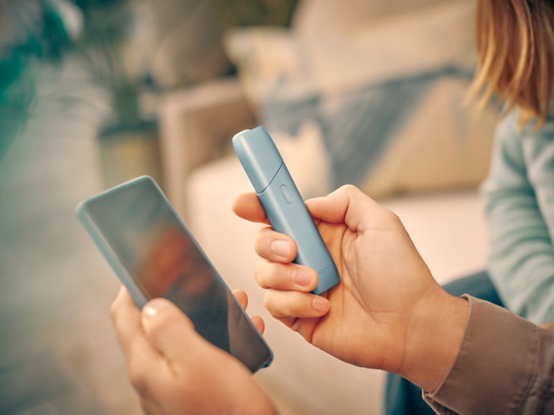 Manos sosteniendo un celular y un IQOS Originals One, mientras inicia sesion en myIQOS para tener la mejor experiencia con IQOS.