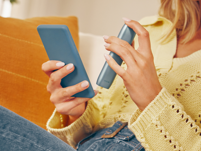 Manos sosteniendo un celular y un IQOS Originals One, mientras descarga IQOS App para realizar un mantenimiento profesional.