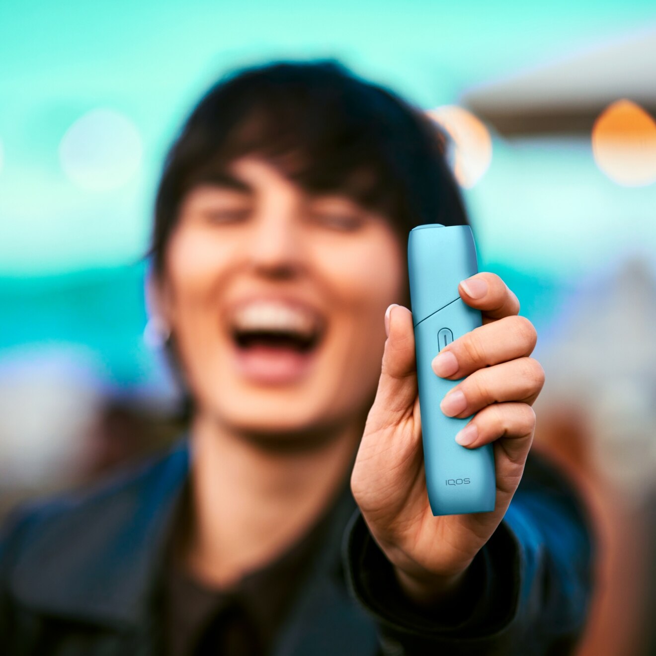 Mujer sonriendo mientras sostiene un IQOS Originals One turquesa. No HEETS